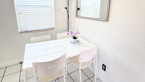 A white dining area with a table, chairs, a mirror, and a window with blinds. - Southwinds 5 for 5 Guests -near the Beach (Hollywood)