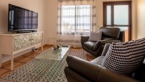 A living room with a flat-screen TV on a white cabinet, a leather sofa with patterned cushions, a glass coffee table, and a patterned rug. - Apartamento Lava in Adeje (Adeje)