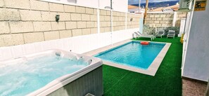 Un bain à remous et une piscine avec une pelouse artificielle, le tout entouré d’une clôture blanche et d’un mur de pierres.