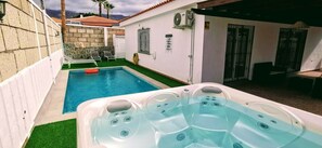 Une piscine avec un jacuzzi, entourée d’un mur blanc et d’un patio couvert.