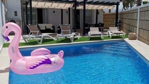 Μια πισίνα με ένα φουσκωτό φλαμίνγκο ροζ χρώματος, ξαπλώστρες και μια σκεπαστή βεράντα.