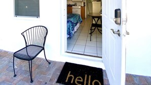 Een voordeur met een deurmat waarop 'hello' staat, een nummer '3' bordje, een stoel en een uitzicht op een kamer met een bed en een tafel.