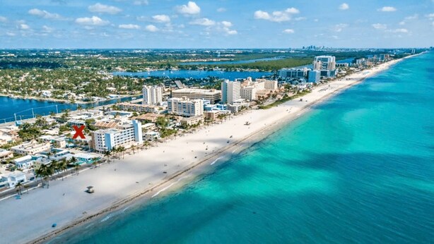 An aerial view of a coastal city with a long sandy beach, clear turquoise waters, and a dense cluster of hotels and buildings along the shore.