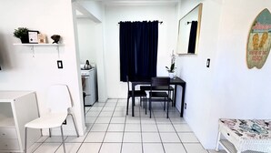 A compact kitchen with a stove, a small table, and a chair. There is a dark curtain, a white wall, and a small shelf with items.