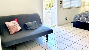 A room with a grey sofa, a bed with patterned bedding, a fish artwork on the wall, and a window with blinds.