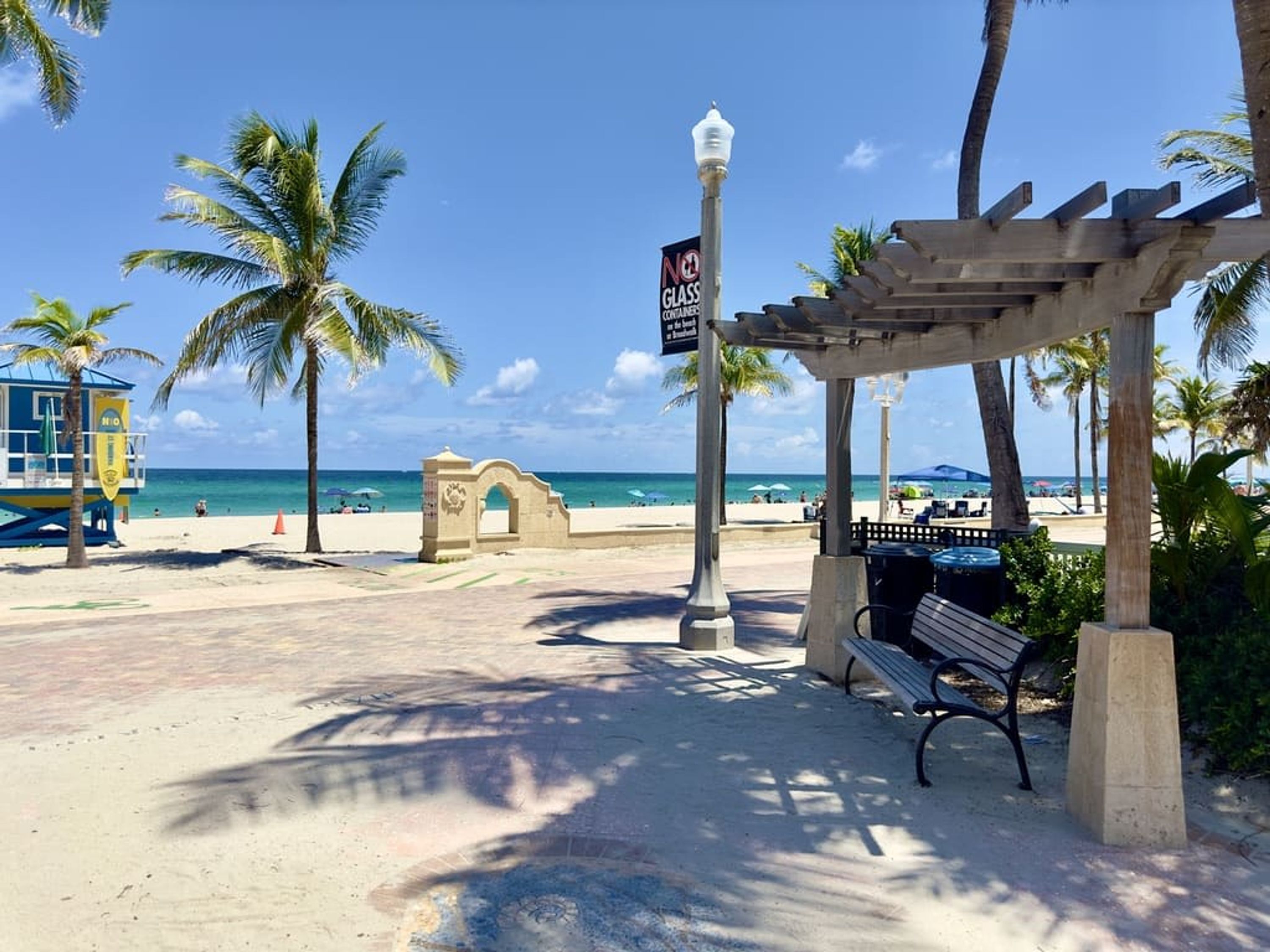 Un paseo marítimo en la playa con una torre de salvavidas, un letrero que prohíbe el vidrio y una pérgola de madera.