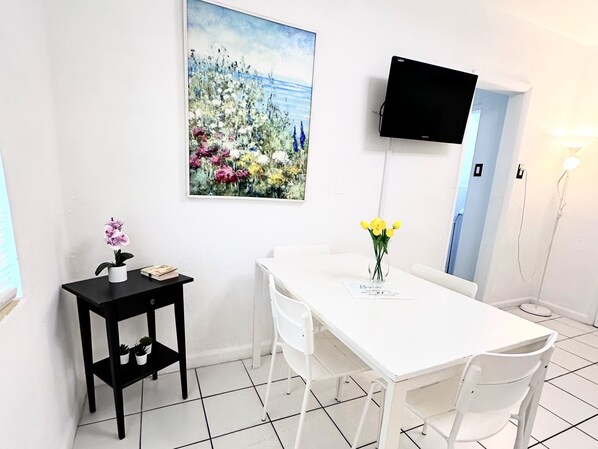 A white dining area with a table, chairs, a black side table with a potted plant, a painting of flowers, and a mounted TV. - Southwinds Inn 7 for 4 Guests Close to the Beach (Hollywood)