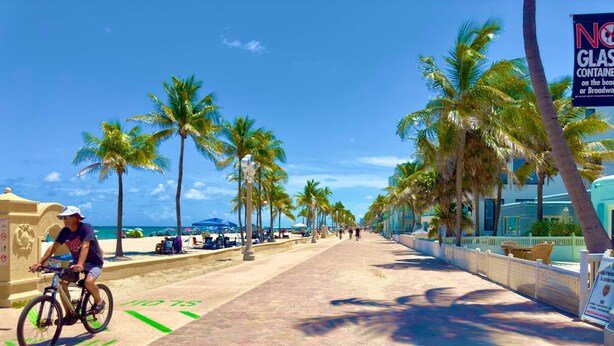 A paved walkway with a cyclist, palm trees, beach umbrellas, and a sign prohibiting glass containers. - Southwinds 8 for 2 Steps to Beach Boardwalk (Hollywood)