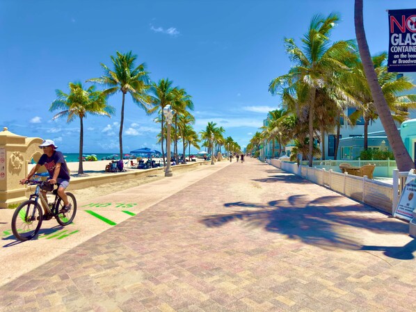Une promenade dallée bordée de palmiers, un cycliste, des parasols de plage et un panneau interdisant l’utilisation de récipients en verre.