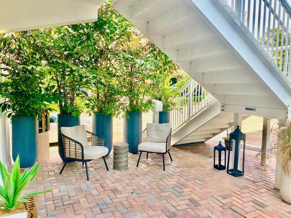 A covered outdoor seating area with brick flooring, two white cushioned chairs, and potted plants. - Southwinds 8 for 2 Steps to Beach Boardwalk (Hollywood)