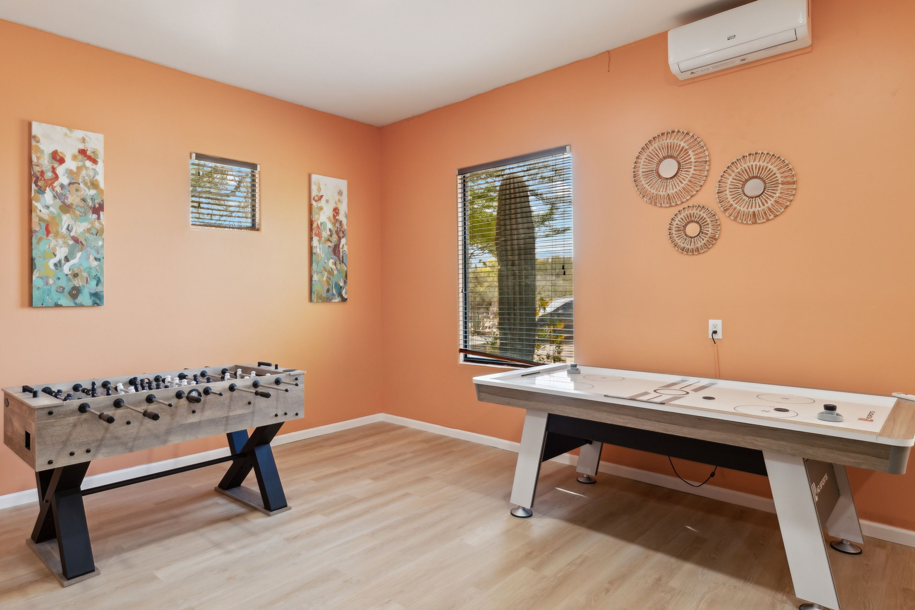 A room with an orange wall, wooden flooring, a foosball table, a pool table, and two windows with blinds.