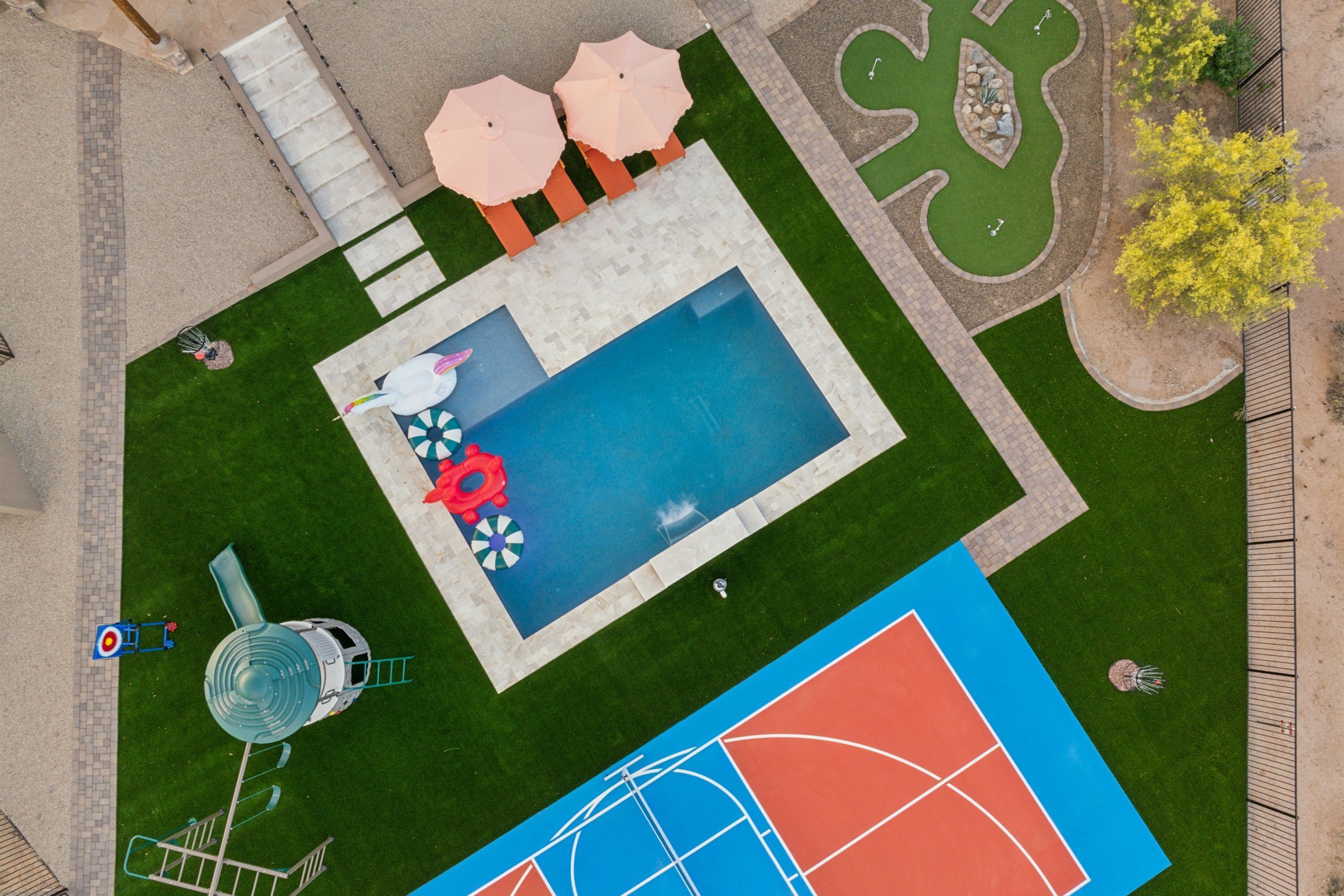 Eine Vogelperspektive auf einen Swimmingpool, der von einem Garten, einem Spielbereich für Kinder und einem Tennisplatz umgeben ist.