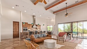 A spacious living area with a large sofa, a round coffee table, and a dining table with chairs. There is a kitchen area with wooden cabinets and a stone fireplace. The room has large windows and a ceiling fan.