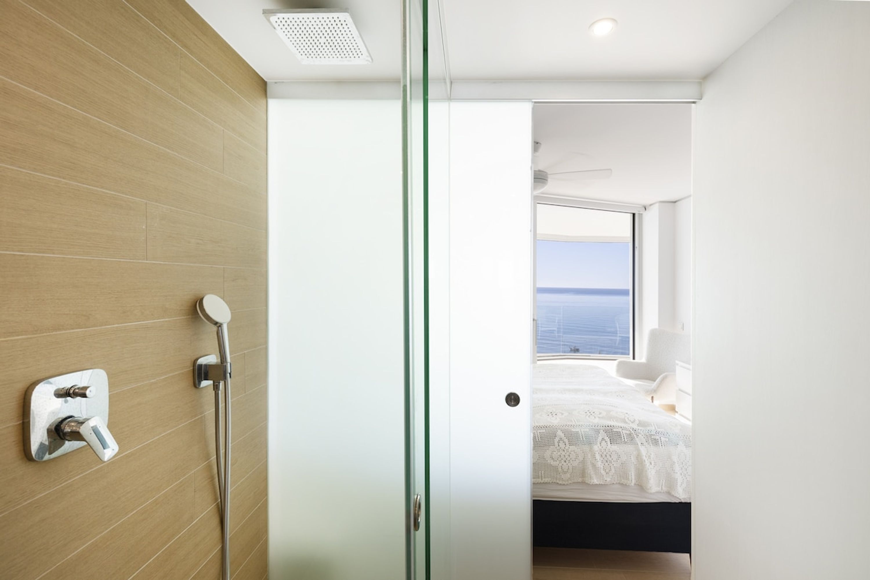 A modern bathroom with a glass shower enclosure, a visible toilet, and a bed with a view of the sea in the adjacent room.