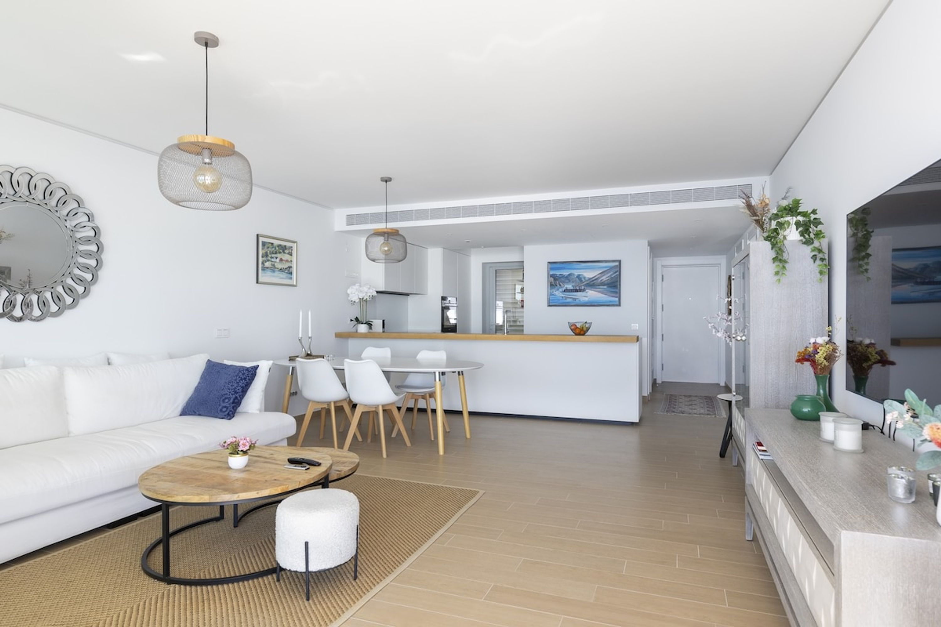 A modern living room with a white sofa, a coffee table, and a dining area with chairs. There is a kitchen in the background with a countertop and a hanging light fixture.