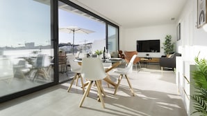 A modern living room with a large glass wall, a dining area with white chairs, a sofa, and a TV mounted on the wall. - Sunset Top Floor Apartment Higueron (Fuengirola)