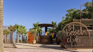 Entree van een strandclub met een pad omzoomd door palmbomen, een centraal zitgedeelte en een bordje met de tekst 'the beach club'.