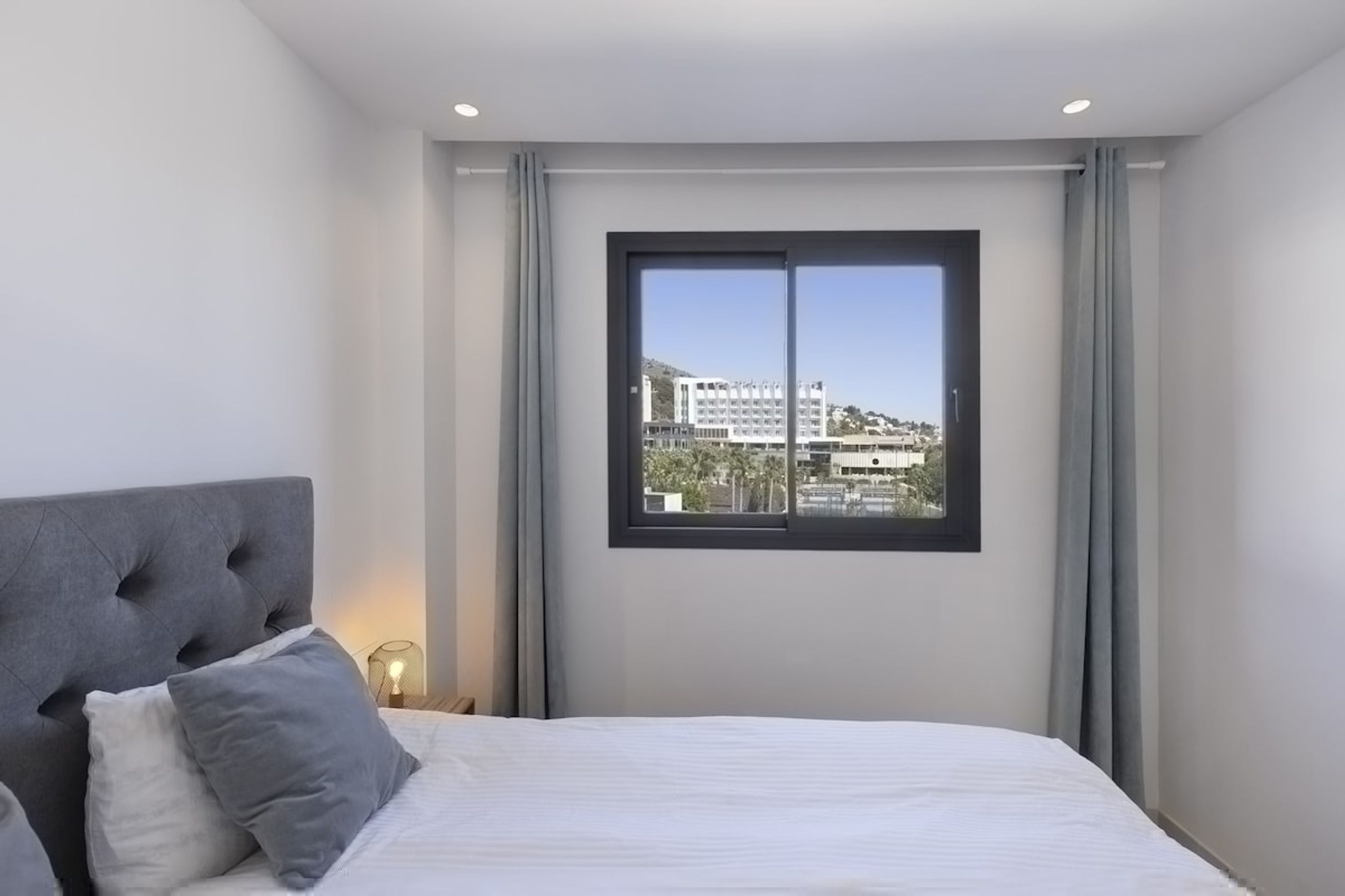 Un dormitorio moderno con una cama grande, cabecera capitonada y vistas a edificios a través de una ventana.