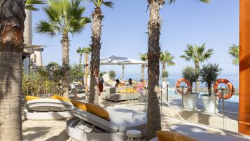 Een zonneterras met ligbedden en parasols, palmbomen en uitzicht op de zee.