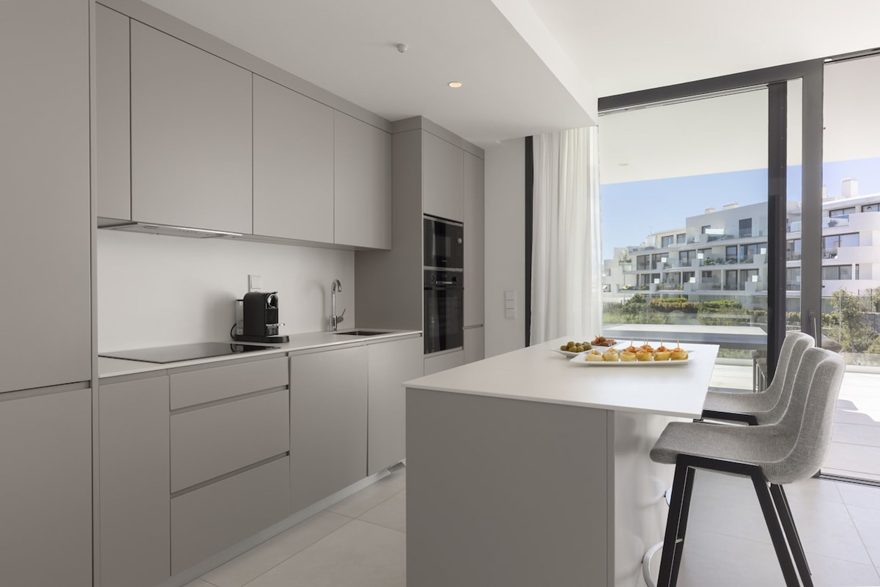 A modern kitchen with a central island, grey cabinetry, and a view of a residential area through large windows.