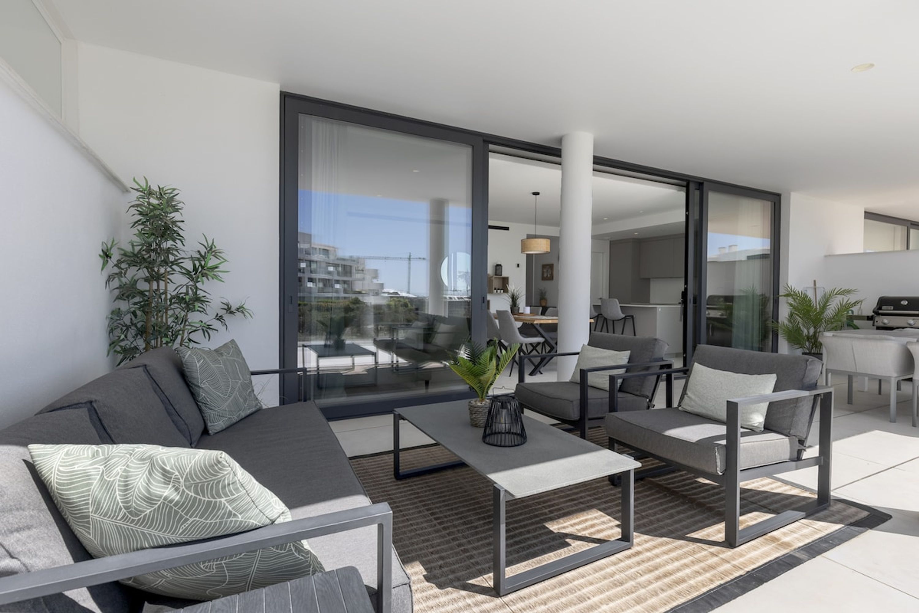 A modern living room with a sofa, coffee table, and chairs, a large window with a view of the city, and a dining area in the background.