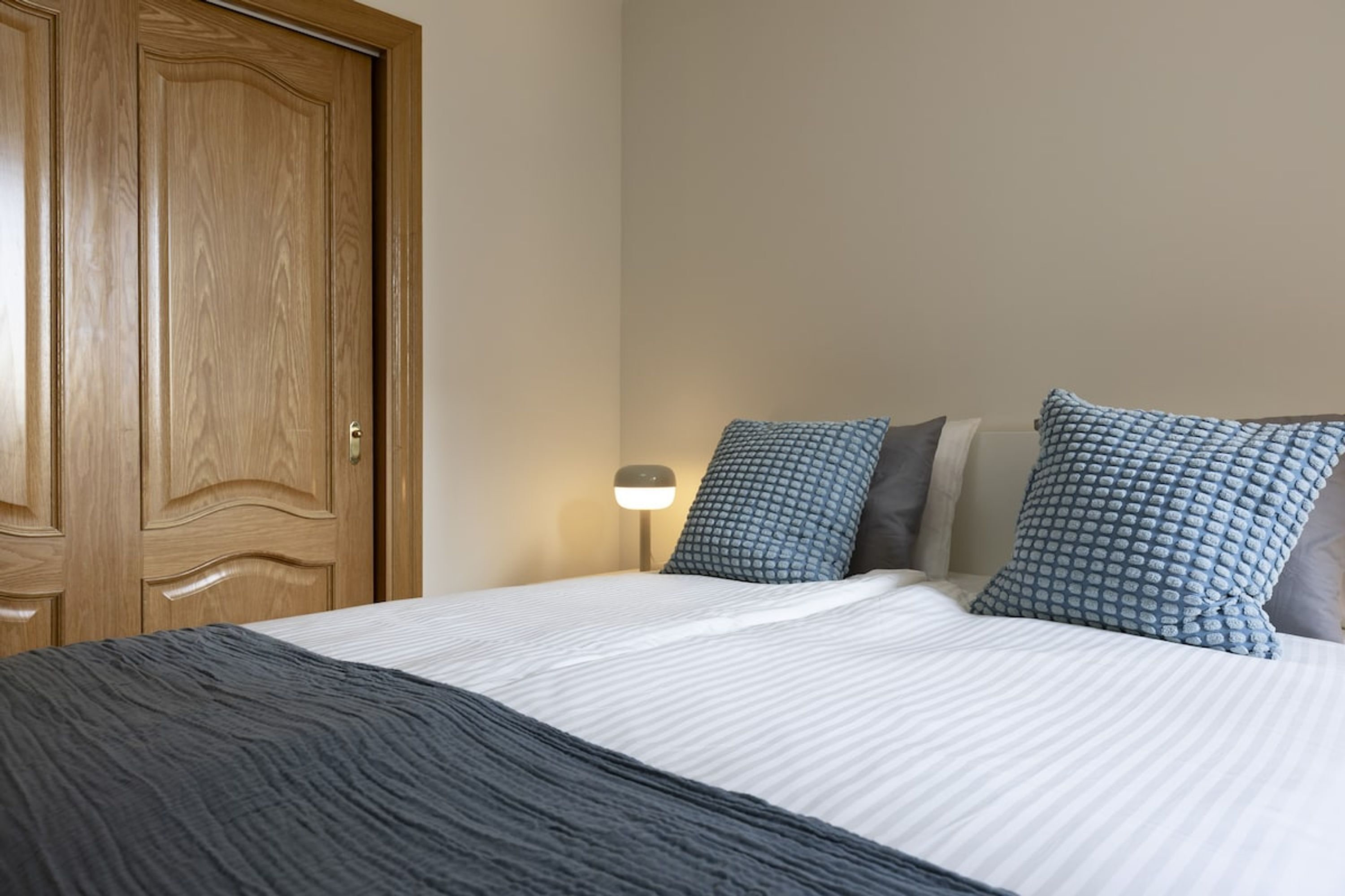 A neatly made bed with a dark blanket and pillows, a wooden door, and a bedside lamp.