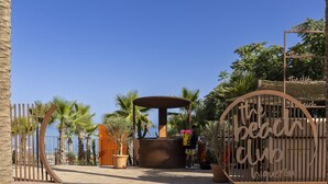 Ingang van een strandclub met een poort, palmbomen en een klein kioskje.