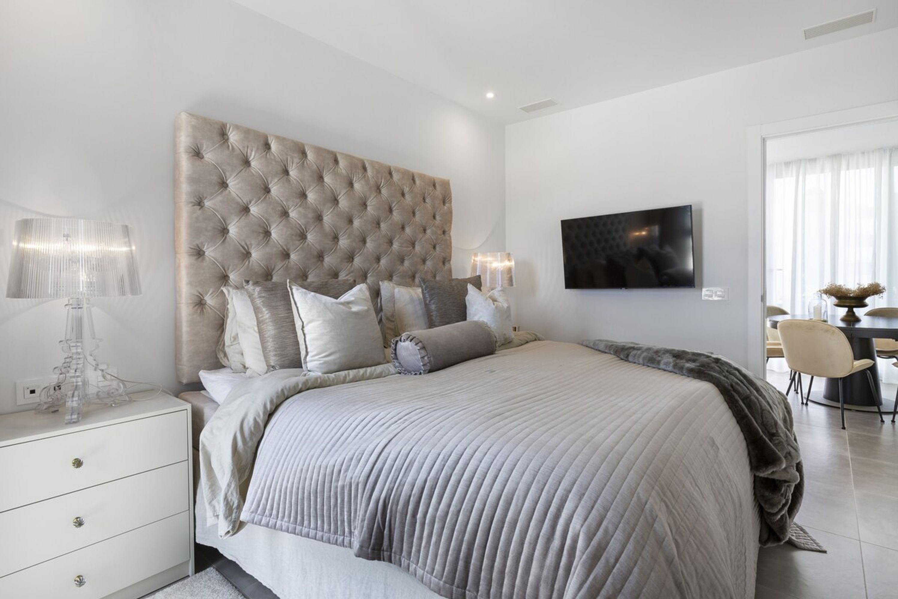 Une chambre moderne avec une tête de lit capitonnée, un grand lit agrémenté de nombreux coussins, une table de chevet avec une lampe et un téléviseur à écran plat fixé au mur.