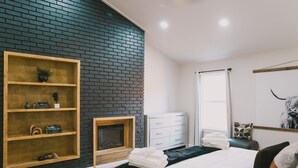 A modern bedroom with a brick accent wall, a wooden shelf with decorative items, a fireplace, a bed with neatly folded towels, a white dresser, and a mirror with a cow head artwork. - Misha: 6443 Redpine Road, Dallas (Dallas)