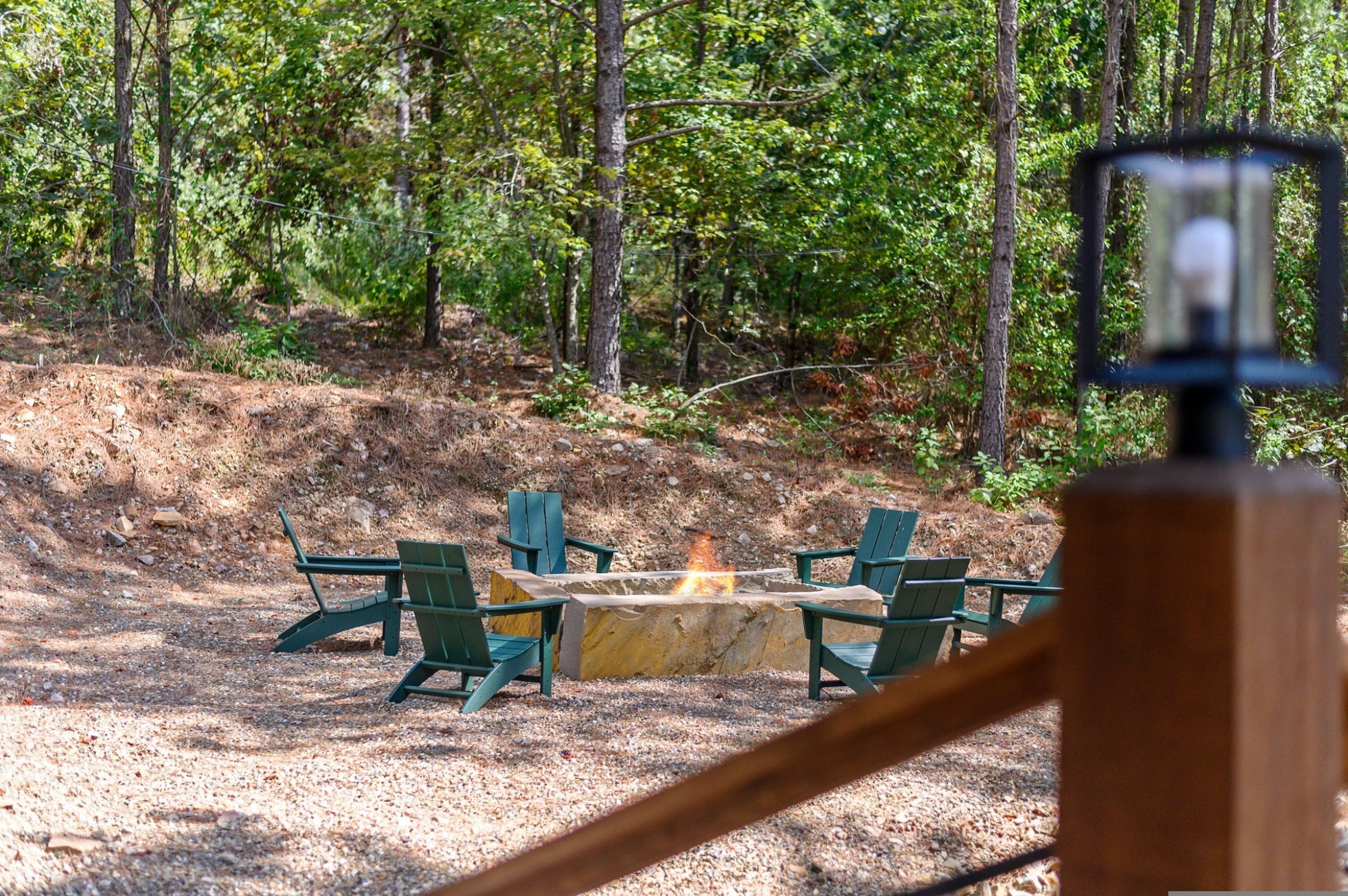 A fire pit with four chairs arranged around it, surrounded by a forested area.