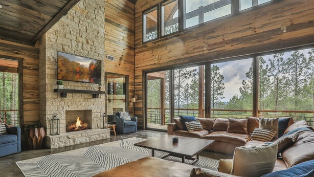 A spacious living room with a stone fireplace, leather sofas, and large windows offering a view of the outdoors. - Timb - Broken Bow - 319 Lukfata Fork Trail (Broken Bow)