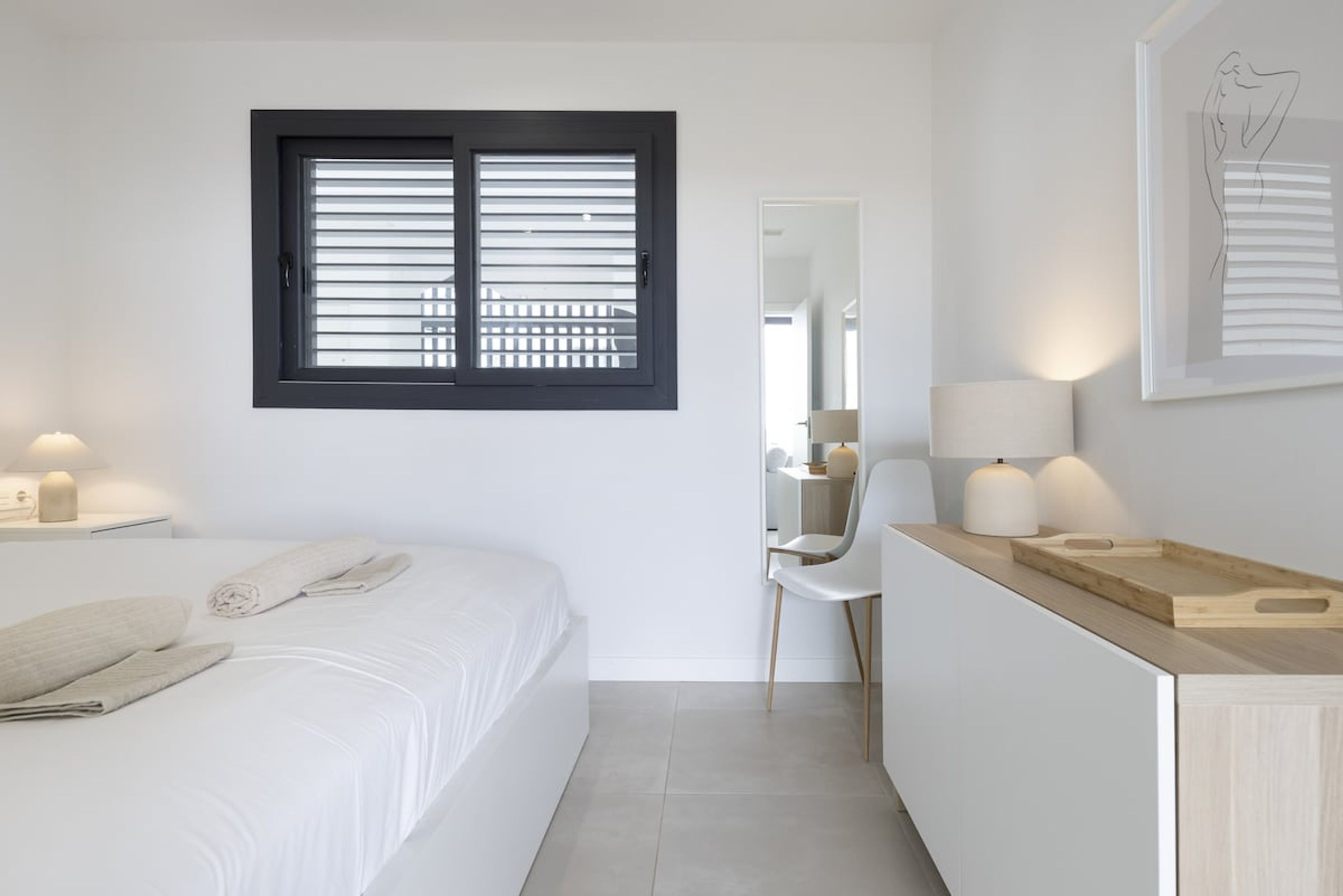 A bedroom with a bed, a bedside table with a lamp, a window with blinds, a mirror, and a wooden side table with a tray.