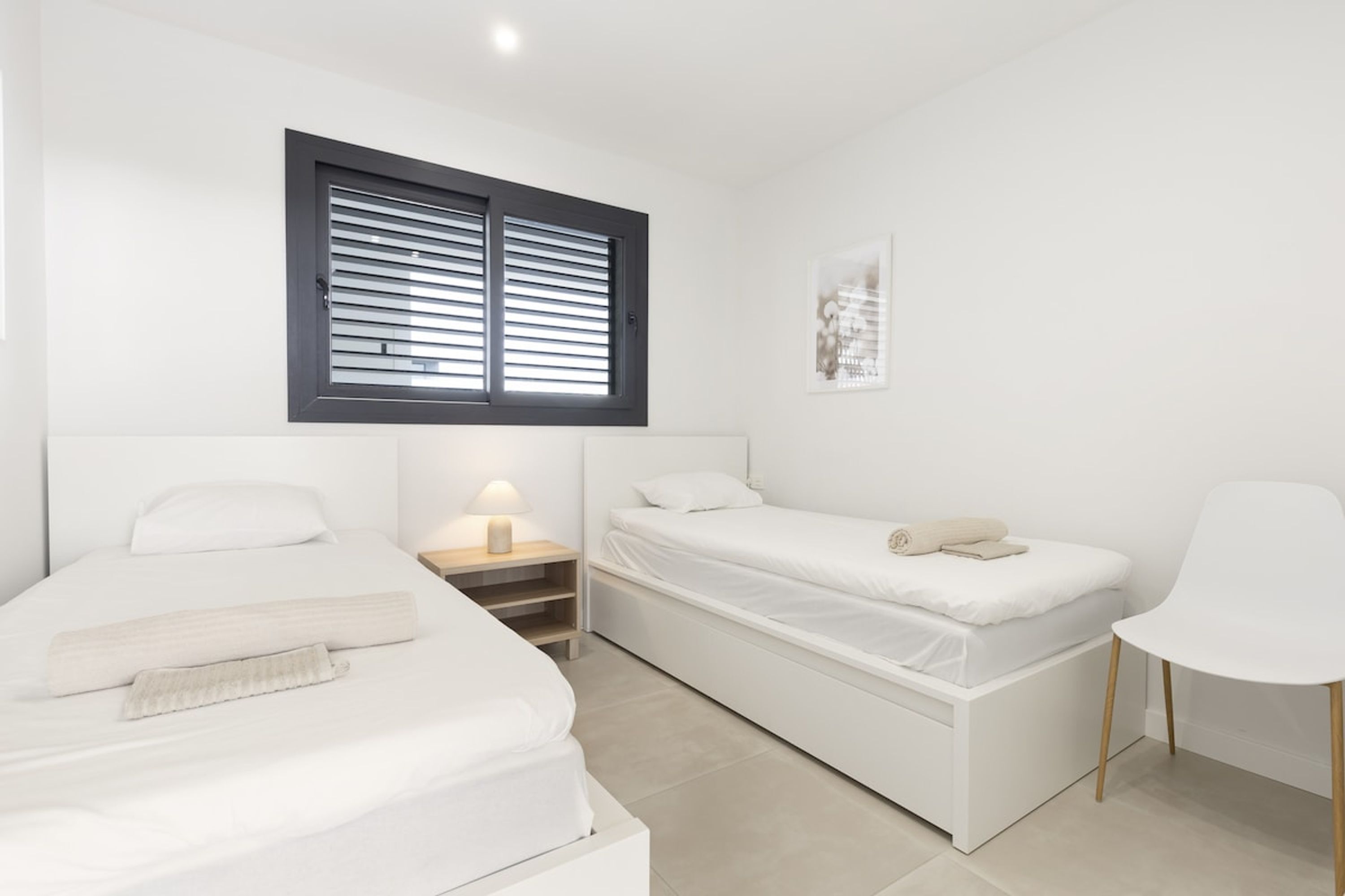 A modern bedroom with a bed, a bedside table with a lamp, a chair, and a window with blinds.