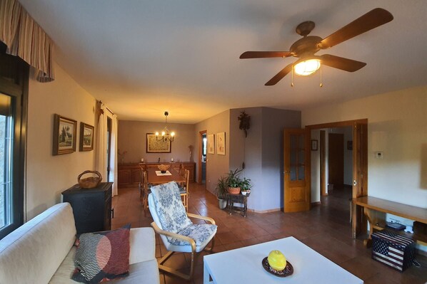 A spacious living room with a sofa, a dining table, and a ceiling fan. There are framed pictures on the walls and a potted plant on a side table. - Edificio Taratola (Ortigosa del Monte)