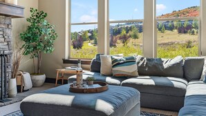 A modern living room with a sectional sofa, a fireplace, a large window with a view of a green landscape, and a small table with a tray. - Nret - Park City - 13546 N Noah Court (Kamas)
