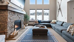 A living room with a stone fireplace, a large flat-screen TV, a gray sectional sofa, and a view of a snowy landscape through large windows. - Nret - Park City - 13546 N Noah Court (Kamas)