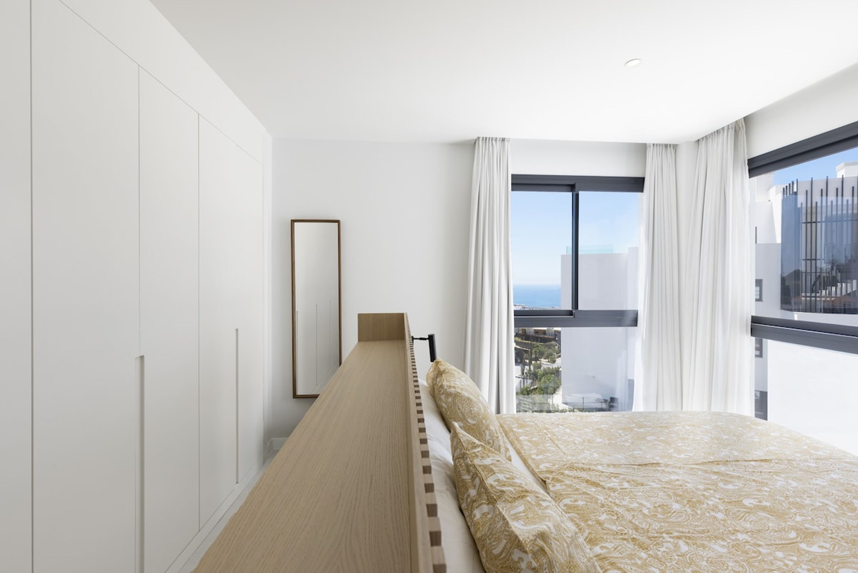 A modern bedroom with a bed, a large window with curtains, and a view of the city.