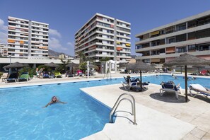 Un espace piscine doté de chaises longues et de parasols, entouré d’immeubles à plusieurs étages.
