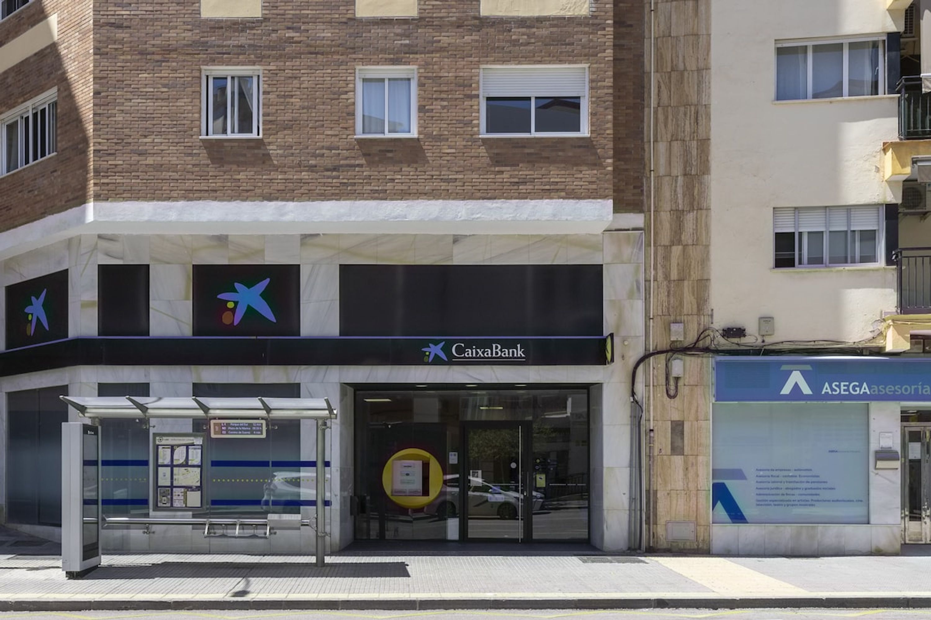 A bank building with a glass entrance, a bus stop, and a sign for CaixaBank.