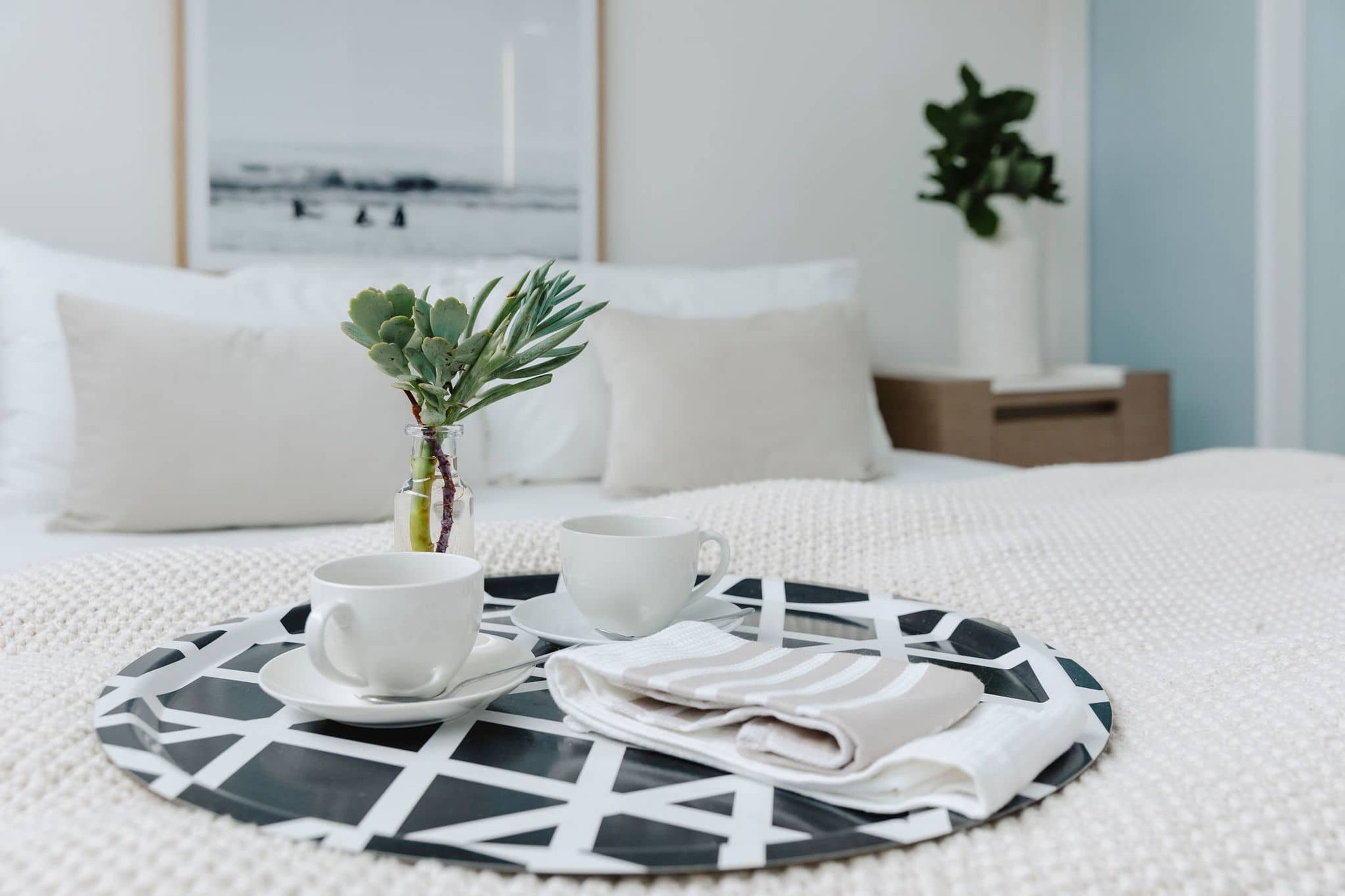 Ein ordentlich bezogenes Bett mit einer weißen Tagesdecke, ein kleiner Topfpflanze, zwei weiße Tassen und ein gefaltetes Handtuch auf einer schwarz-weißen Ablagefläche.