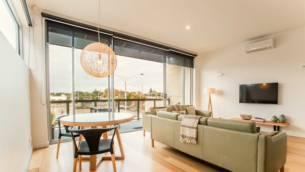 A modern living room with a green sofa, a round wooden table with chairs, a hanging light fixture, and a TV mounted on the wall. - Aquabelle Seven - Couples Retreat That Will Delight (Melbourne)