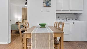 A compact kitchen with white cabinets, a dining table with a green bowl centerpiece, and a bed in the adjacent room. - Flint B (Waco)