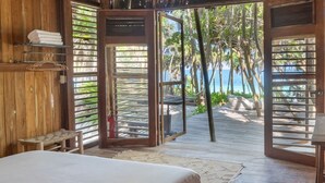 Ein Zimmer aus Holz mit einem Bett, einer Bank und Blick auf Strand und Meer durch offene Türen.