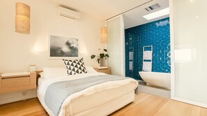 Una camera da letto moderna con un letto grande, comodini, condizionatore d'aria a parete, un quadro incorniciato e un bagno con parete rivestita in mosaico blu e una vasca da bagno bianca.