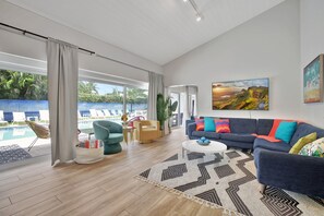 A spacious living room with a large sofa, a coffee table, and a view of a swimming pool. - Seabreeze - 6BR Pool and Steps to Beach (Fort Lauderdale)
