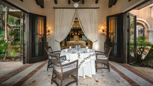 A dining area with a round table set for four, surrounded by wicker chairs, with a view of a kitchen and plants in the background. - Hacienda Palancar Sian Ka an Luxe 11br (Tulum)