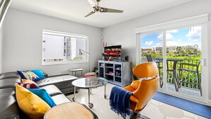 A modern living room with a sofa, armchair, and coffee table, a TV, and a balcony with a view of greenery. - Waterfront Retreat With Pool Steps to Beach (Pompano Beach)
