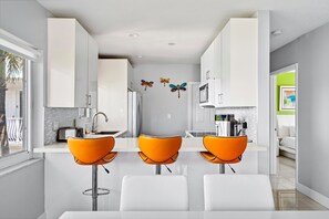 A modern kitchen with white cabinets, a central island with a sink, and three orange bar stools. - Waterfront Retreat With Pool Steps to Beach (Pompano Beach)