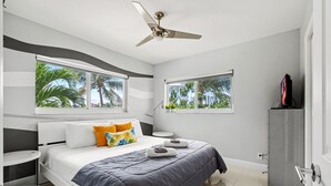 A modern bedroom with a bed, two windows with views of palm trees, a ceiling fan, and a TV on a stand. - Waterfront Retreat With Pool Steps to Beach (Pompano Beach)