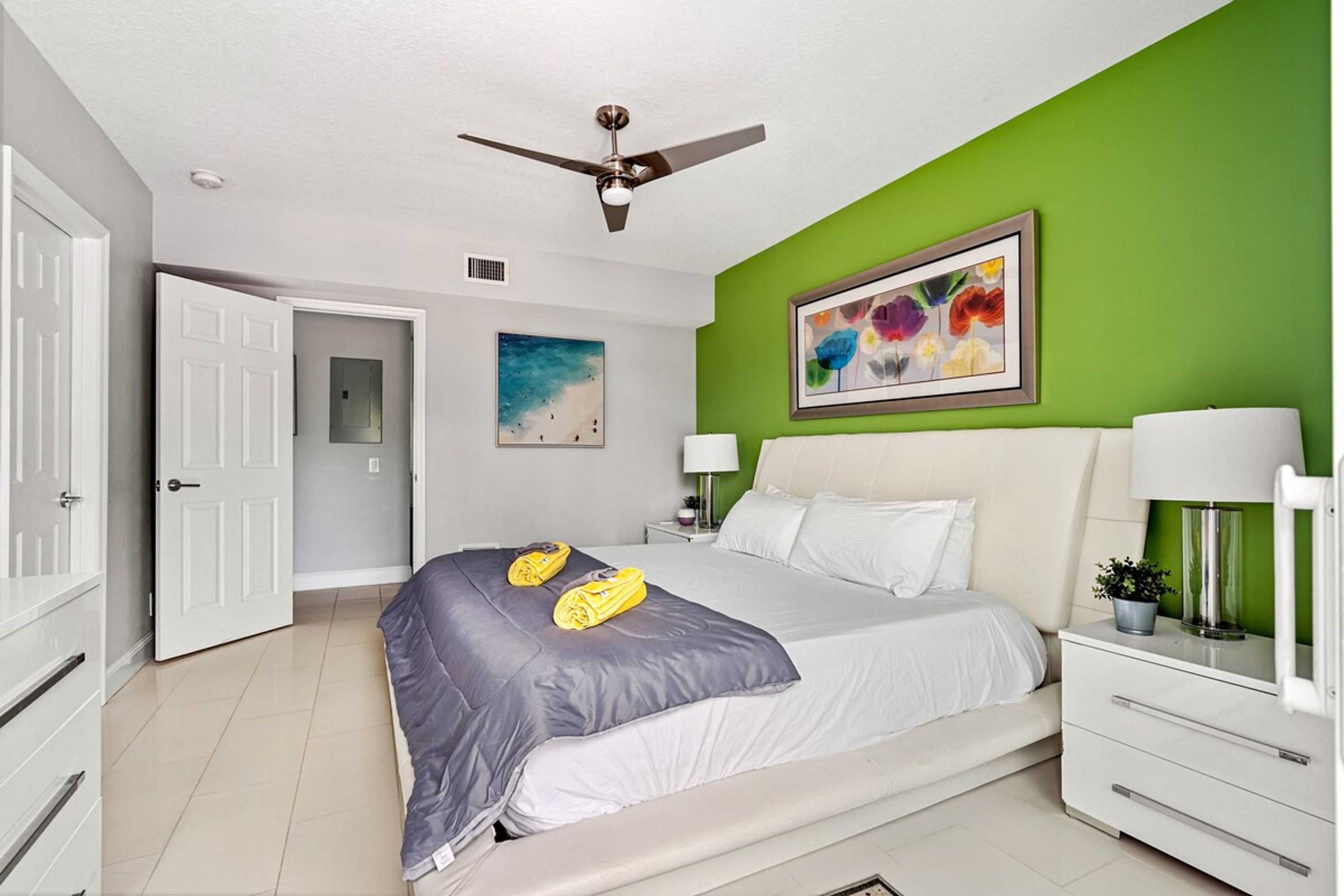 A bedroom with a bed, a ceiling fan, a green wall, a framed painting, and a nightstand with a lamp.